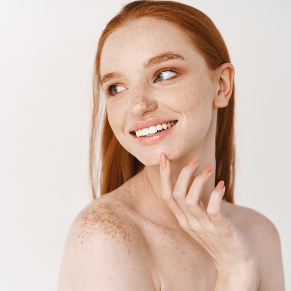 Skincare and beauty. Close-up of young woman with pale skin and freckles standing naked on white background, turn left, smiling white teeth and touching perfect no acne face.