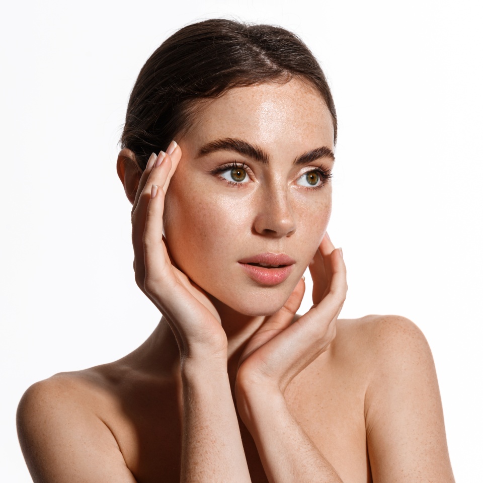 Young beautiful woman with glowing facial skin, touching face and looking away sensually, applying moisturizer or cosmetic cream, standing over white background
