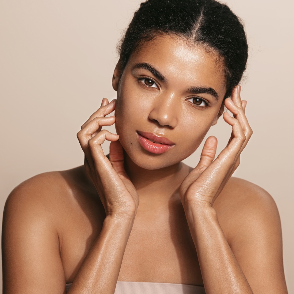 African american woman with clear, glowing facial skin, touches her face with tender gaze, cleansing her body with cosmetic treatment, brown background