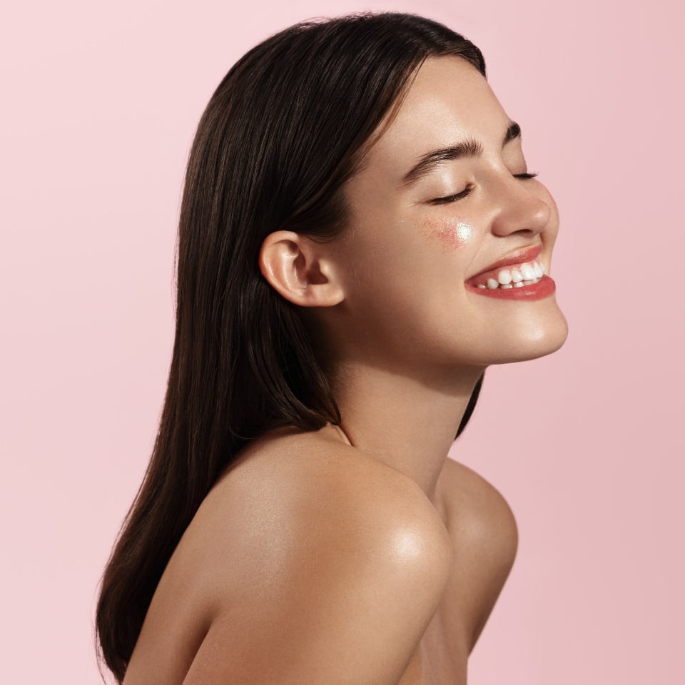 Haircare and beauty concept. Close up of happy smiling woman touches her natural hair, healthy glowing hair, white teeth and clean facial skin, standing against pink background.